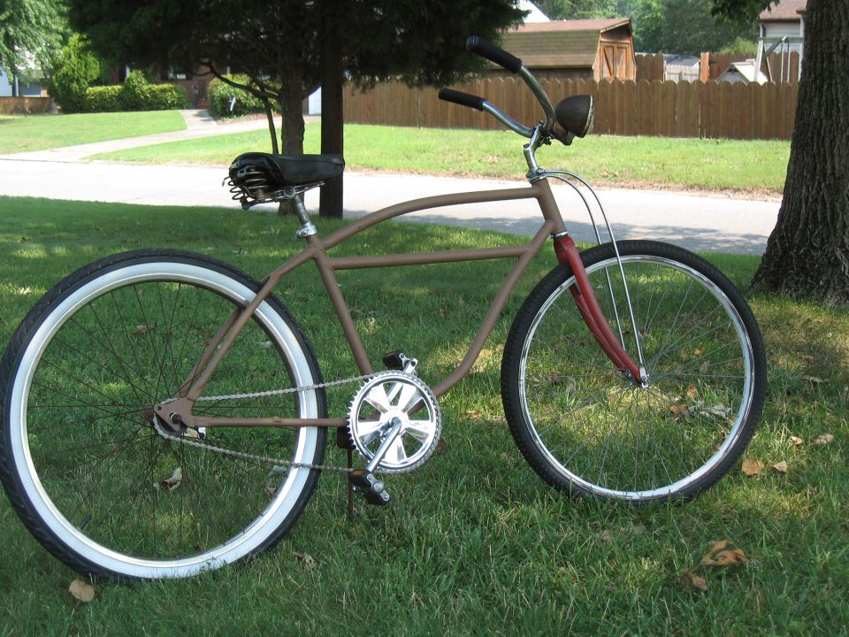 Brown cruiser bicycle with red front fork, black seat, and whitewall tires, parked on grass