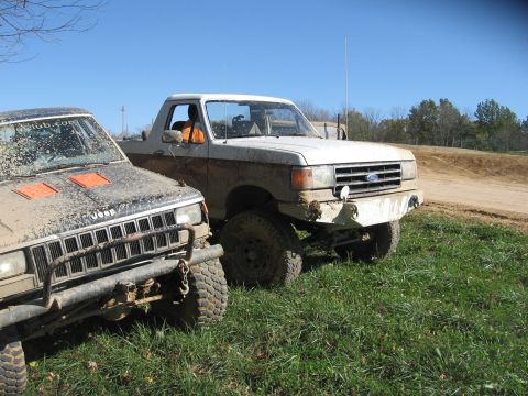 1987 Jeep Comanche - Two off-road vehicles, a Ford Bronco II and a Jeep Cherokee, are parked on a grassy area under a clear blue sky.