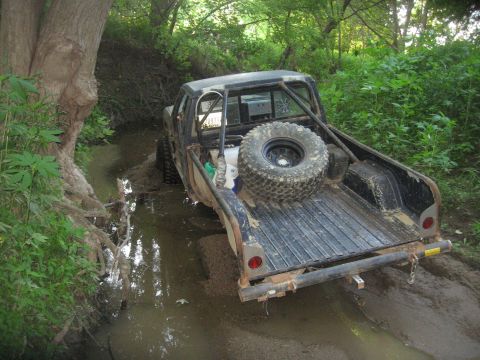 1987 Jeep Comanche - A modified pickup truck navigates a water-filled ditch surrounded by dense foliage.