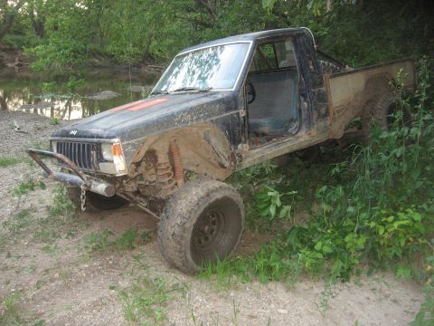 1987 Jeep Comanche - This 1987 Jeep Comanche pickup truck is heavily modified and covered in mud, parked beside a body of water.