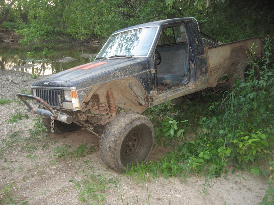 Mud-covered 1987 Jeep Comanche pickup truck with doors removed, parked off-road near water