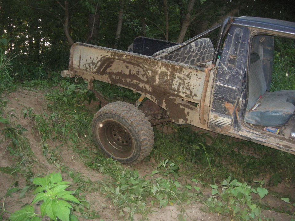 Mud-covered pickup truck with open driver's door, spare tire in bed, parked on a dirt incline