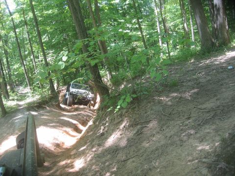 1987 Jeep Comanche - A white Jeep Wrangler navigates a narrow, rutted dirt path surrounded by dense trees.