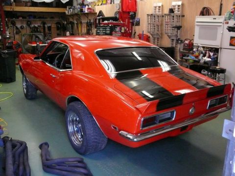 1968 Chevrolet Camaro - A red 1968 Chevrolet Camaro with black racing stripes sits in a garage, with exhaust headers visible in the foreground.