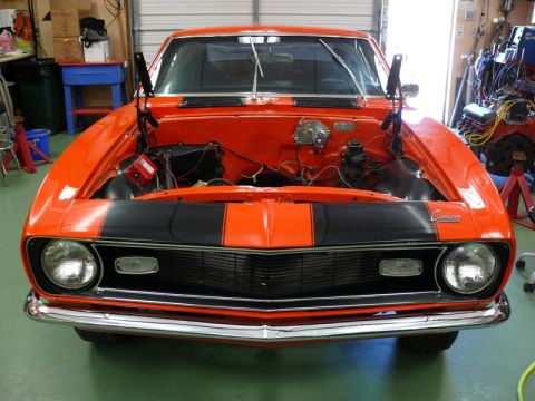 1968 Chevrolet Camaro - Front view of an orange 1968 Chevrolet Camaro with black racing stripes, hood open, in a garage.