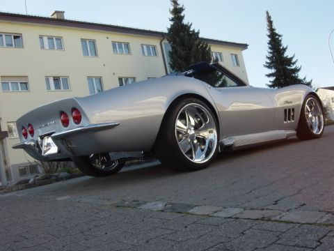1976 Chevrolet Corvette - Silver convertible sports car with custom chrome wheels and side pipes, seen from the rear.
