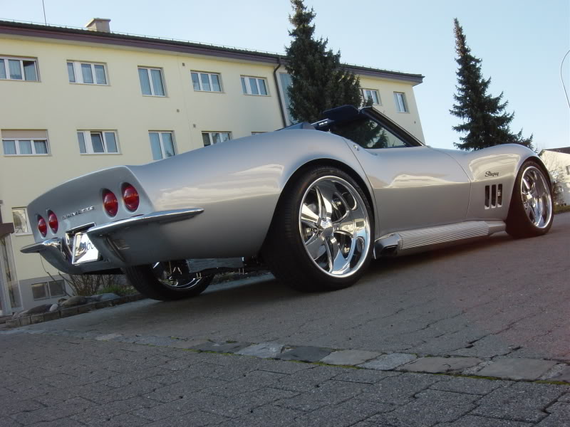 Silver convertible sports car with chrome wheels, parked on a street. Rear view shows quad taillights and side pipes
