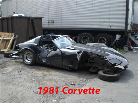 1976 Chevrolet Corvette - This heavily damaged black Corvette, identified as a 1981 model, sits in a yard with a large trailer in the background.