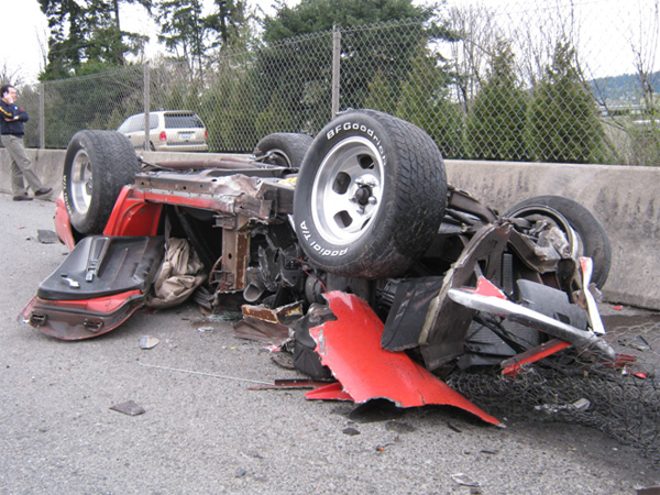 Overturned red sports car with BFGoodrich tires, showing extensive damage and a white SUV in the background