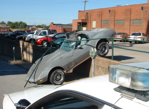 1976 Chevrolet Corvette - A damaged gray Chevrolet Corvette is precariously positioned on a concrete barrier, with a police car visible in the foreground.