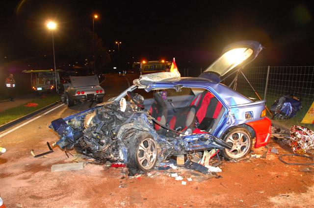 Heavily damaged blue hatchback with red rear fenders, trunk open. Another car, possibly a silver coupe, is in the background