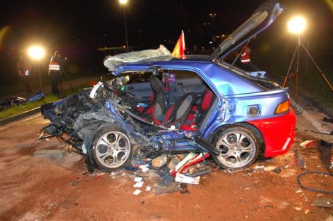 1976 Chevrolet Corvette - A severely damaged blue hatchback with a red rear bumper rests at a crash scene, with emergency lights illuminating the wreckage.