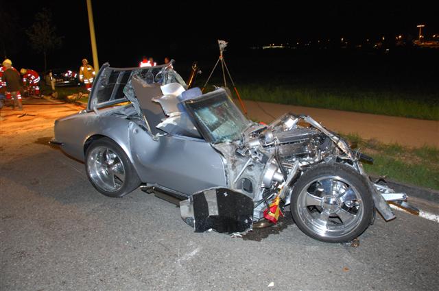 Silver convertible with front end smashed in, roof is damaged, firefighters are in the background