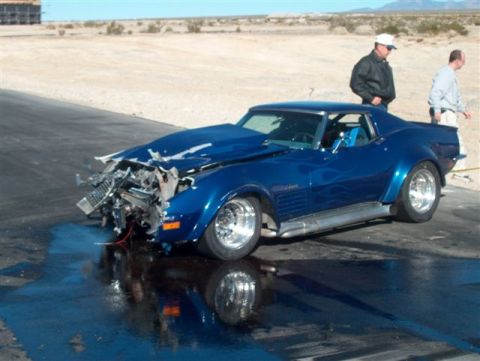 1976 Chevrolet Corvette - A heavily damaged blue Chevrolet Corvette sits on a track, its front end crushed, with two men observing.
