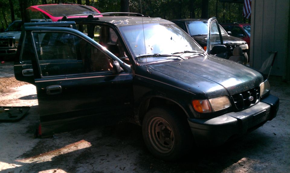 Dark Kia Sportage SUV, front 3/4 view, driver door and hood open on dirt lot