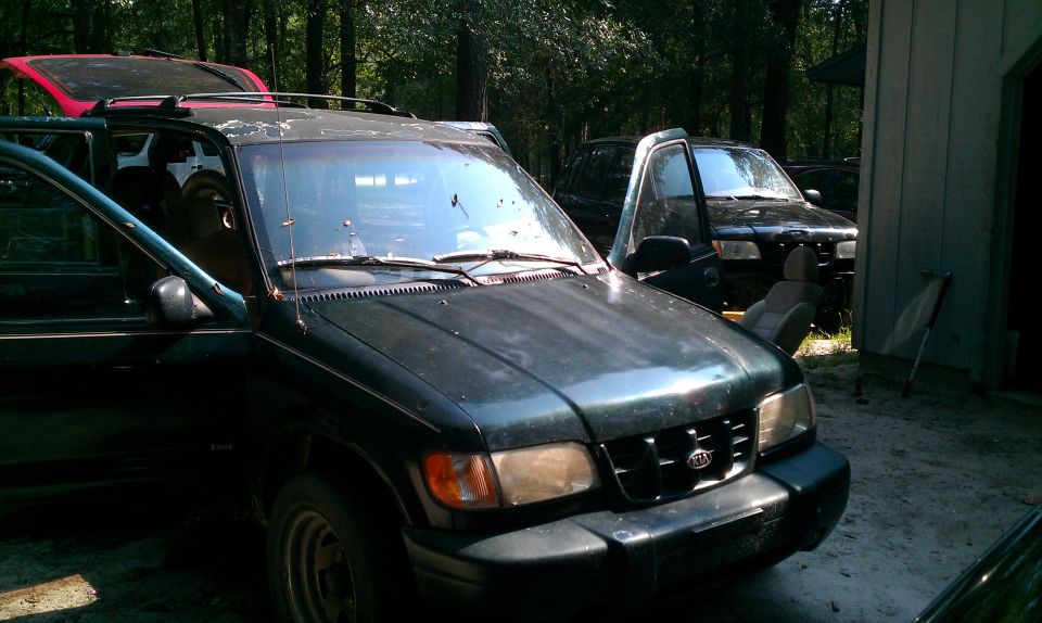 Dusty dark green Kia Sportage SUV, front 3/4 view, driver's door open, in wooded lot