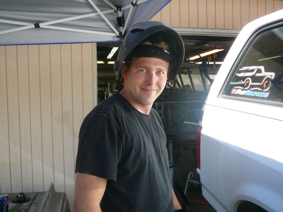 Man wearing a welding mask smiles for the camera next to the rear of a white SUV