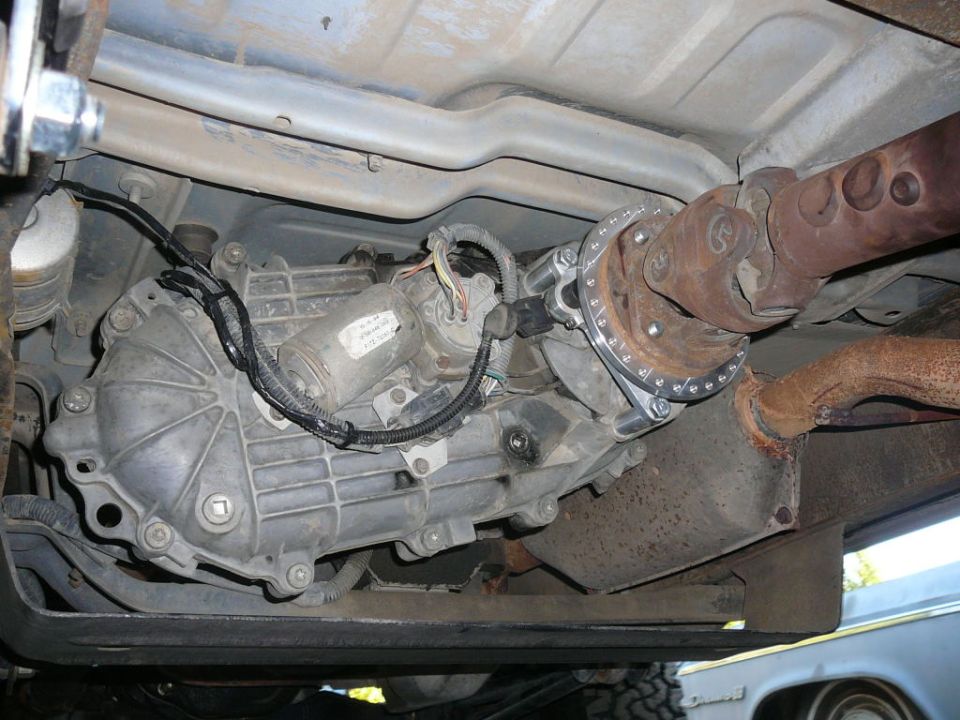 Underside view of a vehicle's transfer case, driveshaft, and exhaust system. Text reads "SUPERMOTORS"