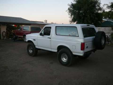 1988 Ford Bronco - White Ford Bronco parked outside with a red Bronco under a carport in the background.