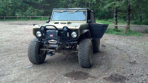1974 Ford Bronco - Olive green 1974 Ford Bronco with open driver's door and winch is parked on a gravel surface in a wooded area.