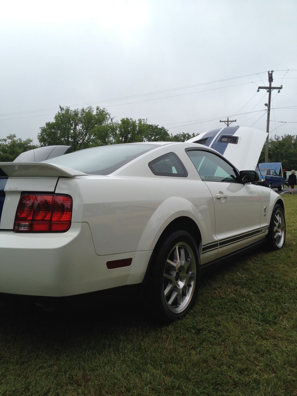 White 2007 Ford Mustang coupe with blue racing stripes on hood and trunk, hood open. Another blue vehicle is visible behind it