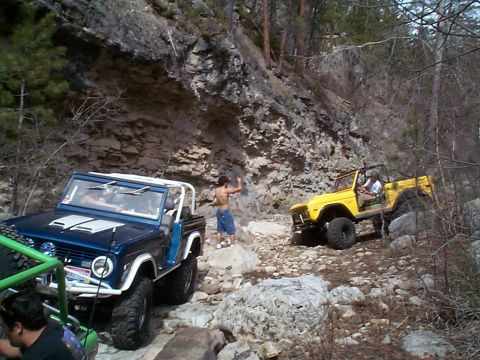 1977 Ford Bronco - Two Broncos navigate a challenging rocky trail in a wooded area.