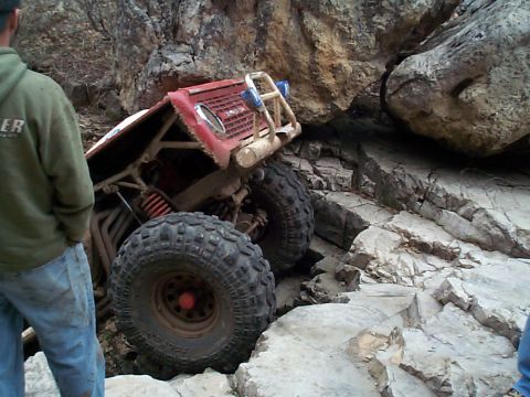 1977 Ford Bronco - A red early Ford Bronco rock crawler tips at a steep angle in a rocky crevice, its front end lifted to show the exposed front axle, red coi…