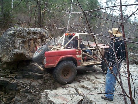 1977 Ford Bronco - A red 1977 Ford Bronco is shown off-roading over large rocks with a person in a cowboy hat observing.