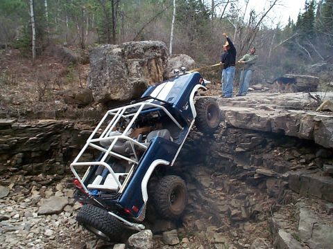 1977 Ford Bronco - Topless dark blue early Ford Bronco with a white exterior roll cage and white fender flares climbing a steep rock ledge at a severe nose-up…