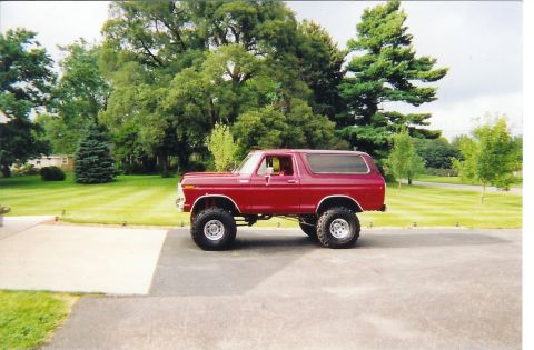 1978 Ford Bronco - A red first-generation full-size Ford Bronco shown in side profile, lifted on large mud-terrain tires with polished wheels, parked on a dri…