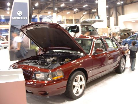 1999 Ford Crown Victoria - A maroon sedan with its hood open is displayed at an auto show, with other vehicles visible in the background.