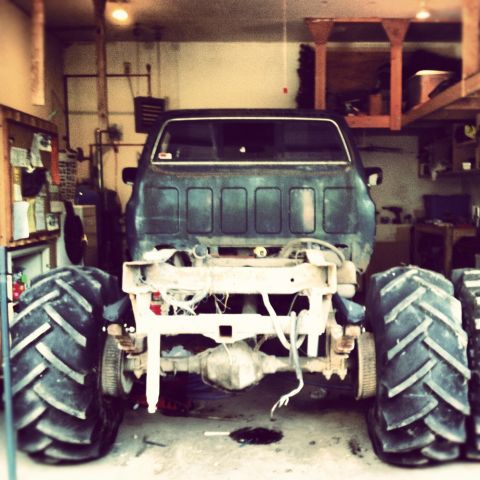 1983 Chevrolet C/K Pickup - Monster truck with oversized tires and a stripped-down rear chassis sits in a garage, awaiting further modifications.