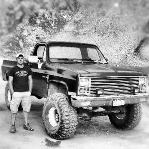1983 Chevrolet C/K Pickup - A man stands beside a modified black pickup truck with large off-road tires.