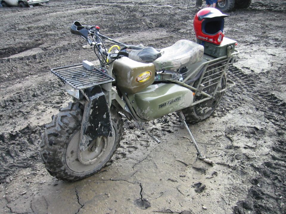 Mud-covered Rokon two-wheel-drive trail motorcycle parked in mud with red helmet on rear rack