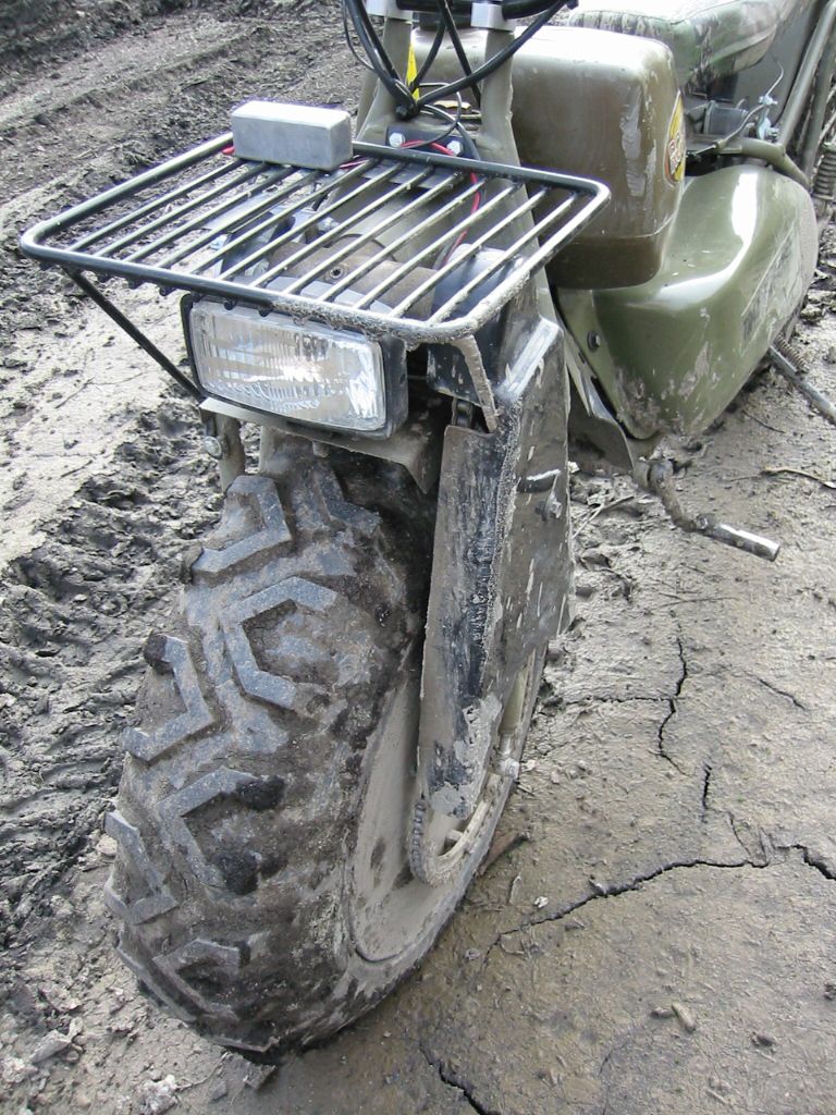 Front of green ATV with tubular rack, headlight and muddy knobby tire on dirt