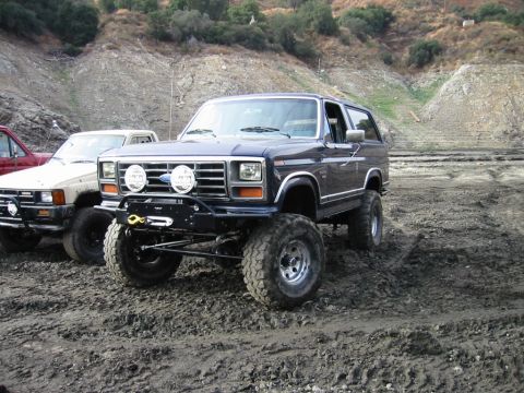 1985 Ford Bronco - A lifted 1985 Ford Bronco with a winch bumper and auxiliary lights sits on muddy ground next to other vehicles.