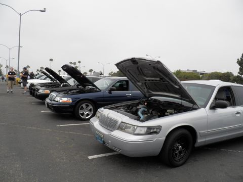 2003 Ford Crown Victoria - Lineup of Ford and Mercury Panther sedans with raised hoods, attendees walking among them.