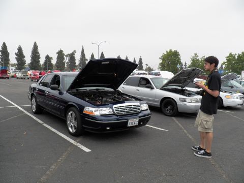 2003 Ford Crown Victoria - Blue Panther sedan with raised hood and BLUE V8 plate as a judge with clipboard inspects it.
