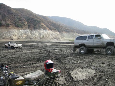 1985 Ford Bronco - Off-road vehicles navigate a challenging muddy terrain with a backdrop of dry, rocky hills.