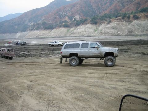 1985 Ford Bronco - A lifted silver SUV with oversized tires drives across a dusty, barren landscape with mountains in the background.