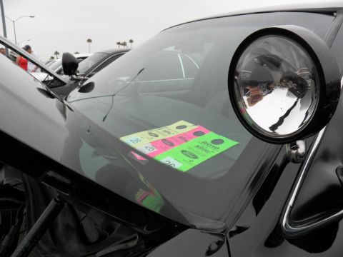 2003 Ford Crown Victoria - Detail of a black car's round headlamp and hood, windshield wearing colorful registration stickers, parking lot behind.