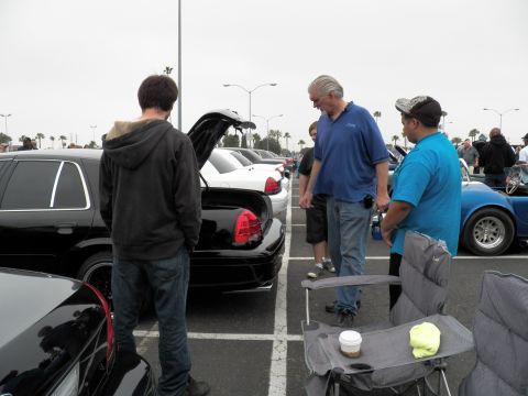 2003 Ford Crown Victoria - Spectators inspect a black sedan's open trunk while folding chairs and a coffee cup sit in the lot foreground.