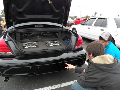 2003 Ford Crown Victoria - Two onlookers crouch to view a black sedan's trunk-mounted subwoofer enclosure with twin speakers and amp.
