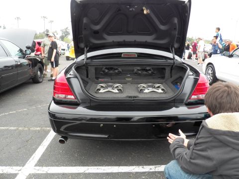 2003 Ford Crown Victoria - Black sedan rear with raised trunk lid revealing a custom dual-subwoofer box; spectators stand nearby.