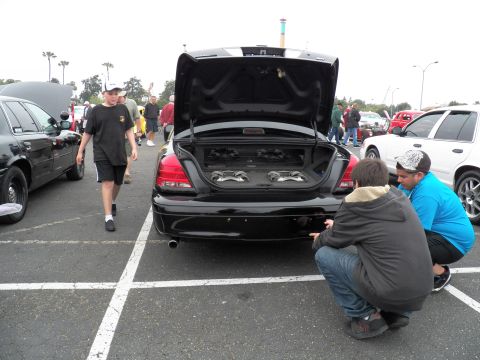2003 Ford Crown Victoria - Rear of a black sedan showing twin trunk subwoofers as two attendees crouch beside it in a parking lot.