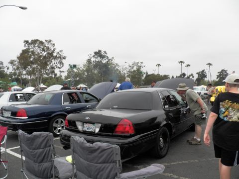 2003 Ford Crown Victoria - Black Ford sedan parked among other cars with hoods up; folding chairs in foreground under overcast sky.