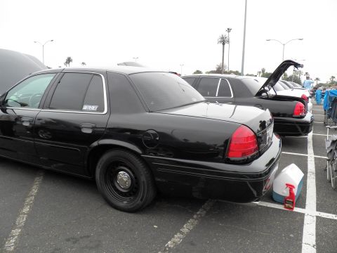 2003 Ford Crown Victoria - Black Ford sedan on plain steel wheels parked in a marked lot, washer jug beside it, overcast sky above.
