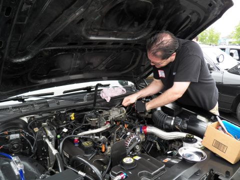 2003 Ford Crown Victoria - Technician bends into an engine compartment, polished intake tube and open parts box nearby, cars behind.