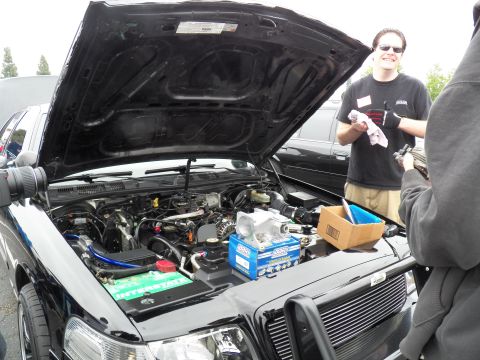 2003 Ford Crown Victoria - Black vehicle's open hood reveals its engine with a blue battery; a grinning man holds a rag beside it.