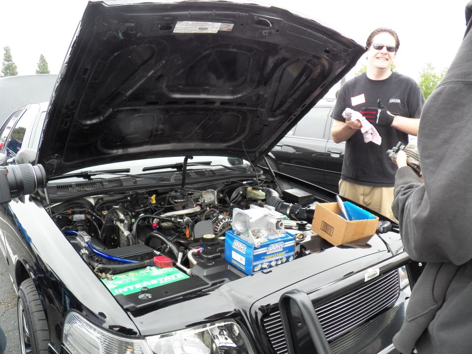 Open hood of black car showing engine with blue battery and intake; smiling man at right